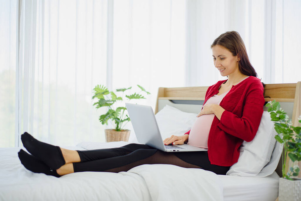 Portrait of pregnant woman in casual home outfit sitting on bed and using laptop with smile and happiness in modern bedroom. - Фото, изображение
