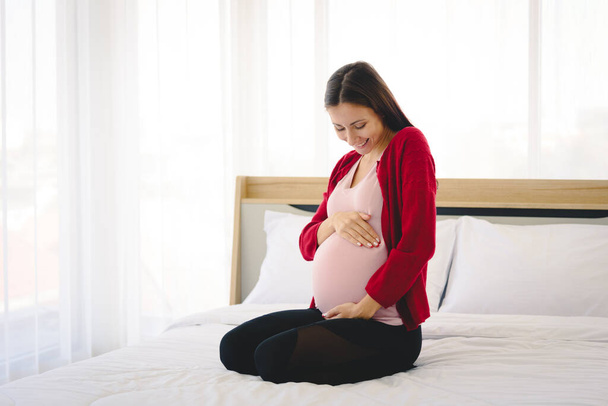 Portrait of pregnant woman in casual home outfit sitting on bed and holding child in her belly in modern bedroom. - Photo, Image