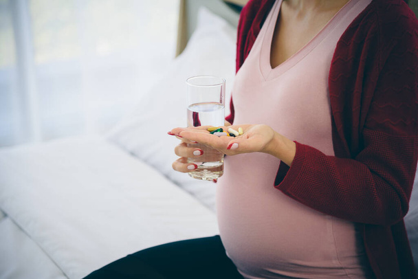 Porträt einer schwangeren Frau im lässigen Heim-Outfit, die auf dem Bett sitzt und im modernen Schlafzimmer ein Glas Wasser und ihre Medikamententabletten in der Hand hält. - Foto, Bild