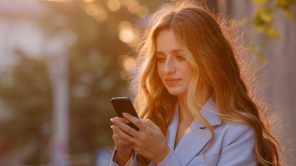 Красивая молодая женщина стоит на улице под открытым небом в солнечных лучах на закате глядя в мобильный телефон отвечая на сообщение онлайн смотреть видео с помощью смартфона чата наслаждаясь интернет Wi-Fi - Фото, изображение