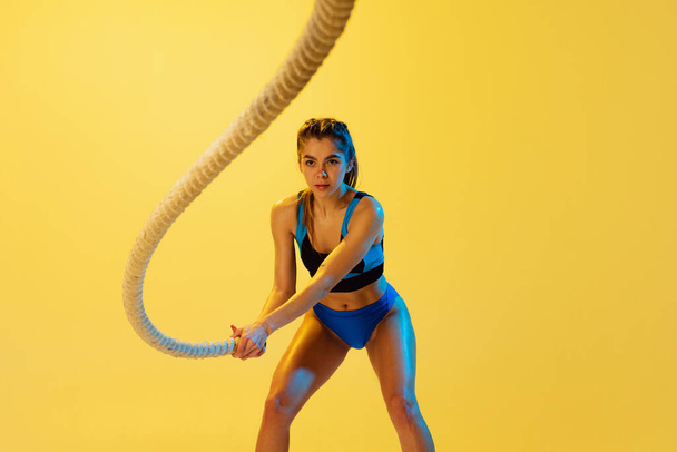 Portrait of young sportive girl training with sports ropes isolated on yellow studio background. Sport, action, fitness, youth and health concept. - Valokuva, kuva