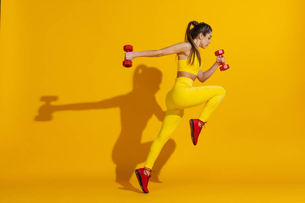 Studio shot of sportive slim girl workout with sports equipment isolated on bright yellow studio background with shadow. Beauty, sport, action, fitness, youth concept. - Zdjęcie, obraz