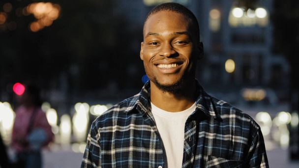 Portrait happy african american 20s millennial satisfied man guy stands in city in evening background looking at camera smiling toothy dental posing outdoors late time male face close up - Photo, Image