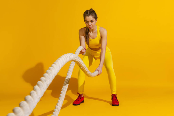 Studio shot of sportive slim girl workout with sports equipment isolated on bright yellow studio background with shadow. Beauty, sport, action, fitness, youth concept. - Foto, imagen