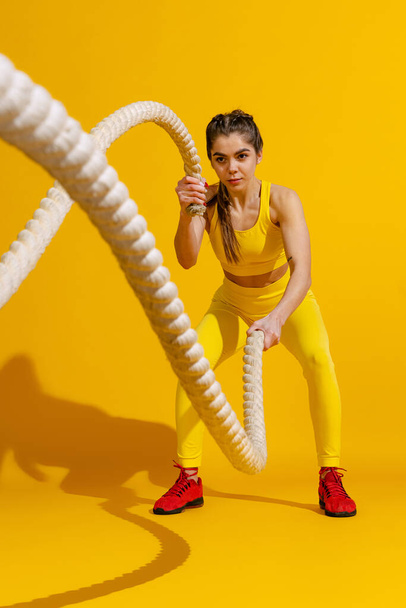 Studio shot of sportive slim girl workout with sports equipment isolated on bright yellow studio background with shadow. Beauty, sport, action, fitness, youth concept. - Фото, зображення