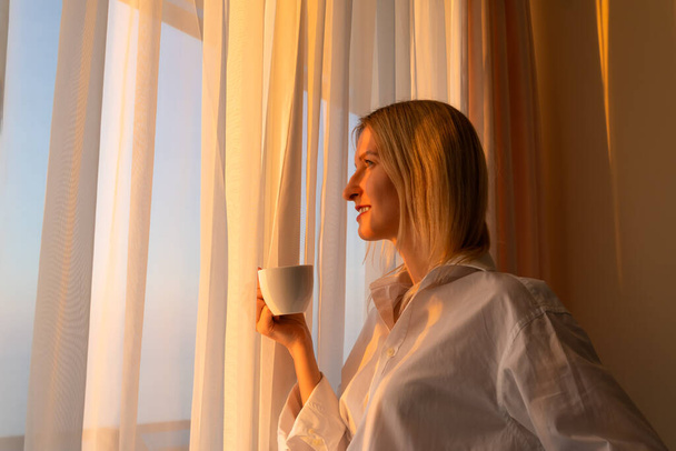 Portret van een jonge glimlachende vrouw in een wit shirt met een witte kop koffie of thee voor het raam en de zonsondergang bewonderend. Begrip geluk, harmonie - Foto, afbeelding