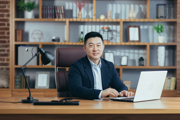 Portrait of a successful Asian businessman, man working in the office sitting at the table, looking at the camera and smiling, happy banker with laptop - Foto, Imagem