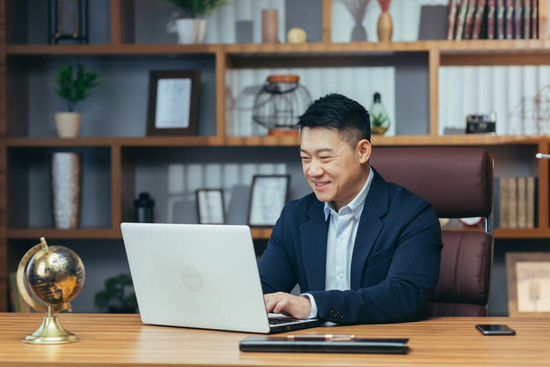 Портрет успішного азіатського бізнесмена в класичному офісі, який працює над ноутбуком, посміхається - Фото, зображення