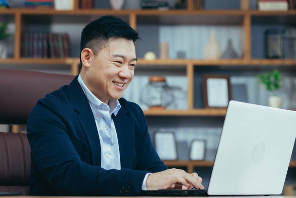 Portrait of a successful Asian businessman in a classic office working on a laptop, smiling - Valokuva, kuva