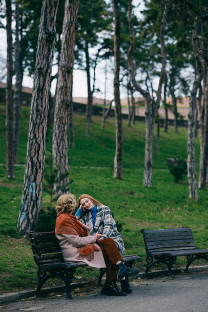 Genç bir kızıl kadın, soğuk havada büyükannesiyle oturuyor ve bankta konuşuyor.. - Fotoğraf, Görsel
