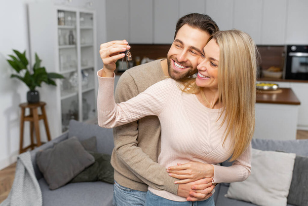 Excited caucasian couple in love move in a new apartment. Cheerful woman in embrace of bearded guy holding the keys - Photo, image