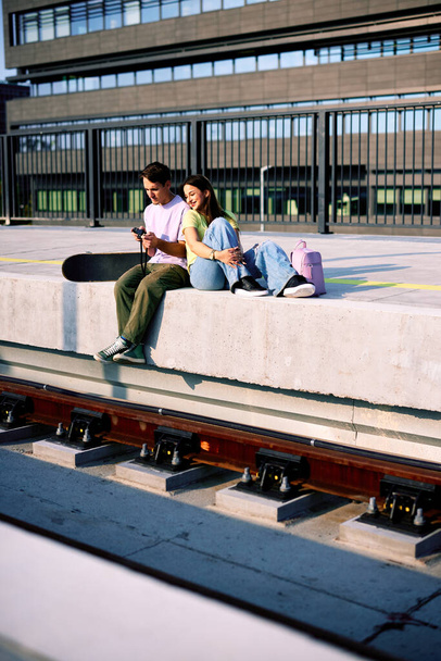 Teenagers are sitting at the railroad station on a sunny day, chilling and using the camera. - Фото, изображение