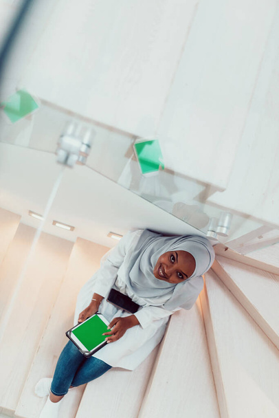mladá africká moderní muslimka pomocí tabletu počítače, zatímco sedí na schodech doma na sobě hidžáb oblečení horní pohled - Fotografie, Obrázek