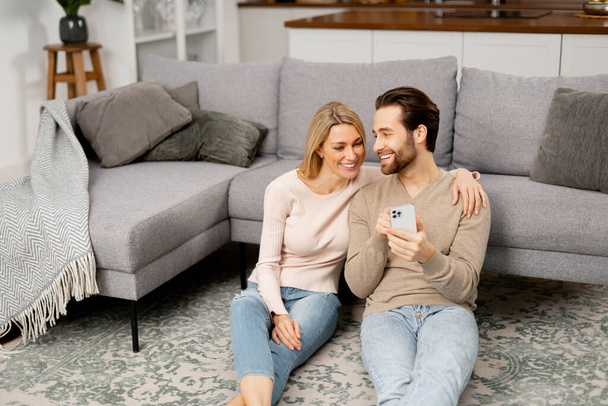 Bearded guy is showing something to his girlfriend. Caucasian couple with smartphone on the floor spending leisure time in social networks, scrolling feed - Foto, imagen