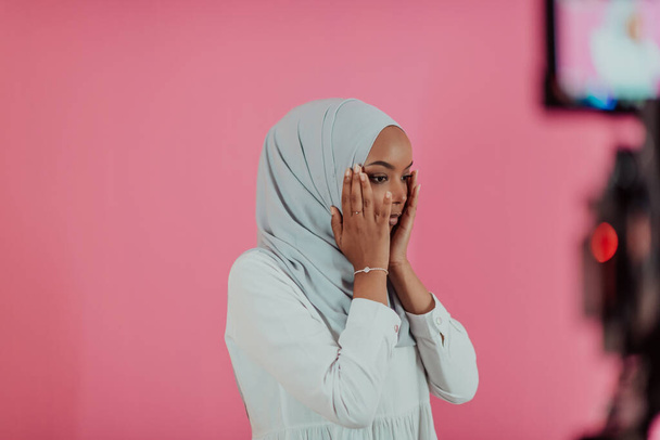 Modern African Muslim woman makes traditional prayer to God, keeps hands in praying gesture, wears traditional white clothes, has serious facial expression, isolated over plastic pink background - Foto, imagen
