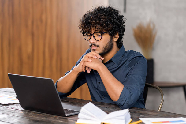 Focused and concerned Indian man in smart casual shirt looking at laptop screen. Serious guy sitting at the desk and resting chin on hands while feeling doubts - Foto, imagen