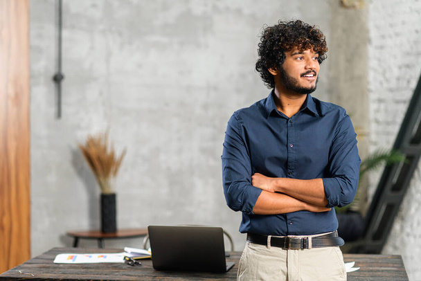 Indian businessman standing in modern office near desk with a laptop and looking away. Ambitious smiling multiethnic man in smart casual wear - 写真・画像