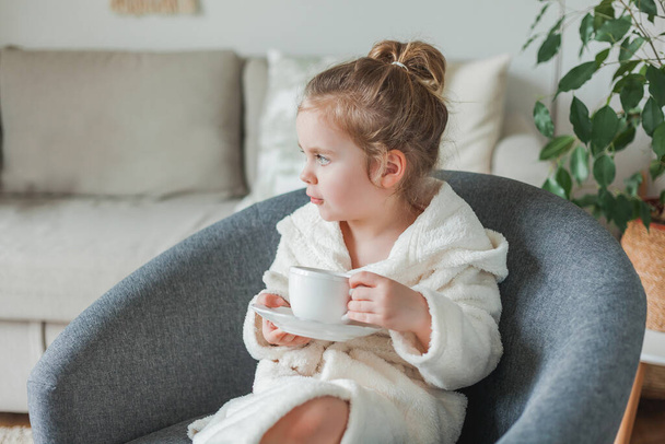 Little curly-haired girl in a soft light dressing gown drinks tea and relaxes at home sitting in a comfortable chair. The baby imitates her mother. Beautiful homely cozy interior. - Photo, image