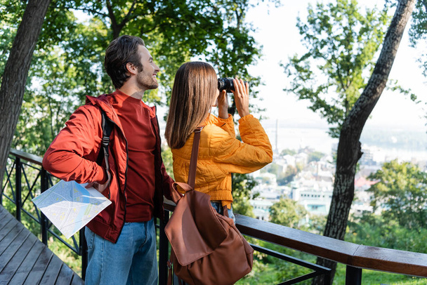 Smiling tourist holding map near girlfriend with binoculars in city  - Photo, Image
