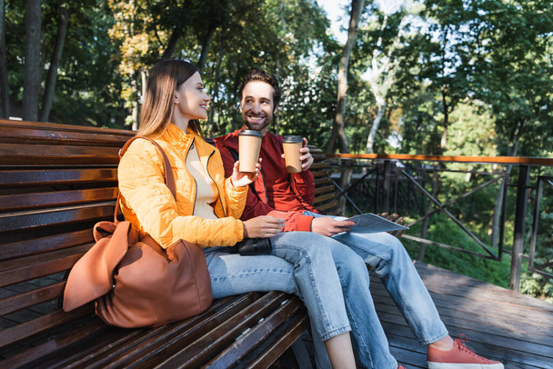 Lächelnde Touristen mit einer Karte, auf der sie Kaffee trinken, um draußen auf der Bank zu sitzen  - Foto, Bild