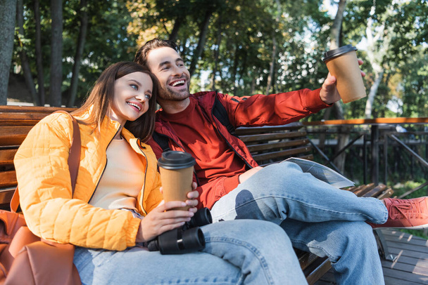 Usmívající se turista drží kávu jít a mapovat poblíž přítelkyně s dalekohledem na lavičce venku  - Fotografie, Obrázek