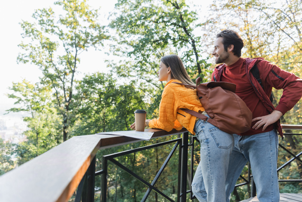 Vue latérale du voyageur souriant regardant loin près de sa petite amie avec sac à dos, café à emporter et carte en plein air  - Photo, image