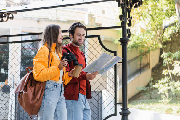 Positive tourist holding map near girlfriend with coffee to go and binoculars on urban street  - 写真・画像