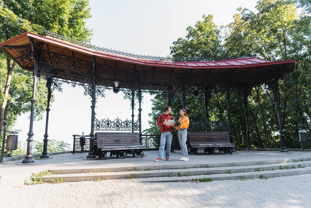 Viajantes sorridentes com café para ir e binóculos conversando com namorado com mapa na rua urbana  - Foto, Imagem
