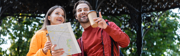 Low angle view of cheerful man holding map and pointing with finger near girlfriend with coffee to go on urban street, banner  - Photo, Image