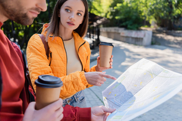 Turysta trzymając kawę iść i wskazując palcem na mapie w pobliżu chłopaka na świeżym powietrzu  - Zdjęcie, obraz