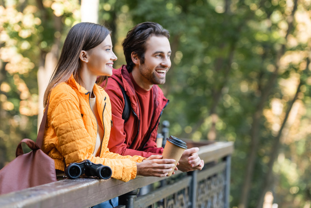 Smiling traveler holding takeaway drink near boyfriend and binoculars on railing outdoors  - Foto, Bild