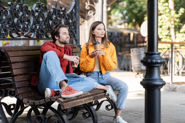 Young tourist holding coffee to go near boyfriend and map on bench outdoors  - Foto, Bild