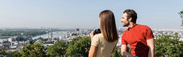 Positive tourist looking away near brunette girlfriend with binoculars on viewpoint in city, banner  - Foto, Bild