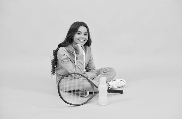 veselé dítě sedět ve sportovním oblečení s badminton raketa a láhev vody na žlutém pozadí, relaxovat - Fotografie, Obrázek