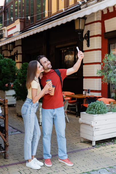 Positive couple with backpacks and coffee to go taking selfie on urban street  - Foto, imagen