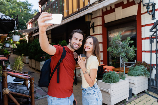 Lächelnder Reisender macht Selfie mit Freundin mit Imbissgetränk auf Stadtstraße  - Foto, Bild