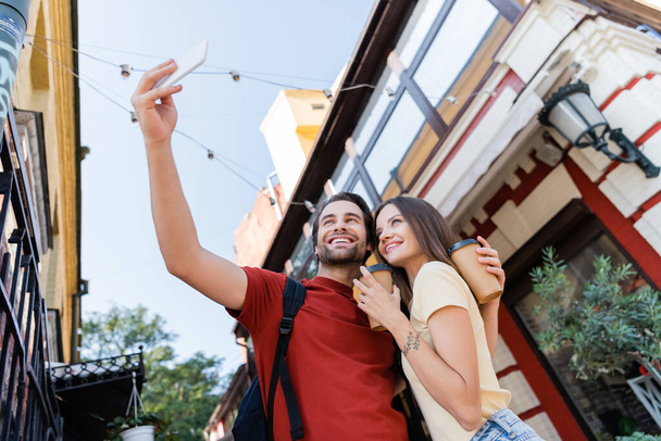 Vista de ángulo bajo de turistas positivos con vasos de papel tomando selfie en la calle urbana  - Foto, Imagen