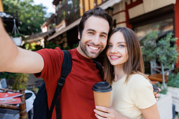 Positive travelers with backpack and coffee to go looking at camera outdoors  - Photo, Image