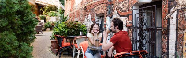 Mujer positiva mirando novio con taza de papel en la terraza de la cafetería, pancarta  - Foto, Imagen