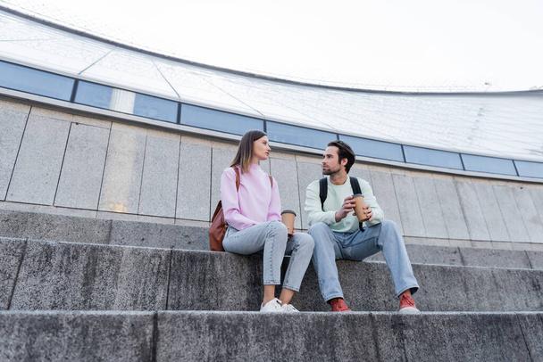Mladí cestovatelé s donáškou pít mluvit na schodech na městské ulici  - Fotografie, Obrázek