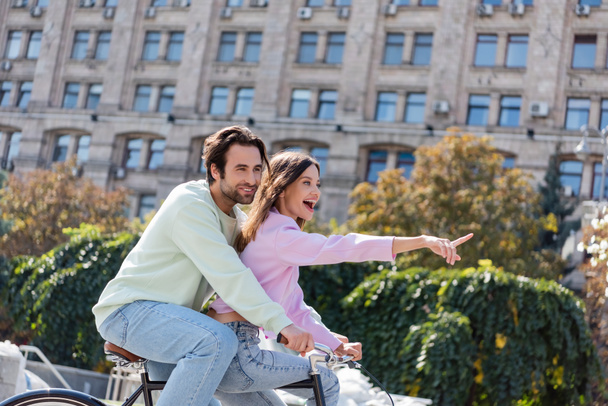 Excited woman pointing with finger while riding bike with boyfriend on urban street  - Foto, Imagen
