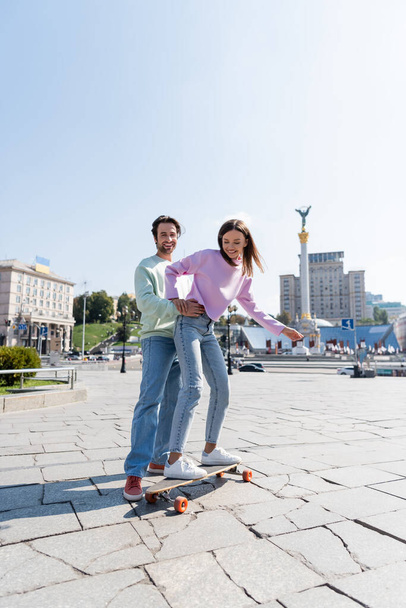 KYIV, UKRAINE - SEPTEMBER 1, 2021: Happy man hugging girlfriend riding penny board on urban street in Kyiv - Foto, afbeelding