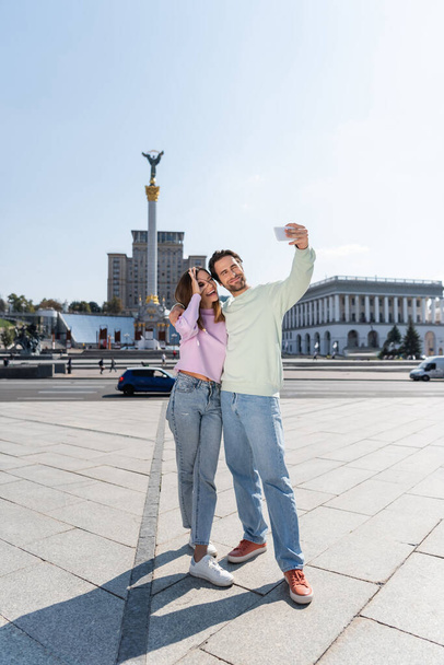 KYIV, UKRAINE - 1 ΣΕΠΤΕΜΒΡΙΟΥ 2021: Χαμογελαστό ζευγάρι που βγάζει selfie στο κινητό του στην Πλατεία Ανεξαρτησίας στο Κίεβο - Φωτογραφία, εικόνα