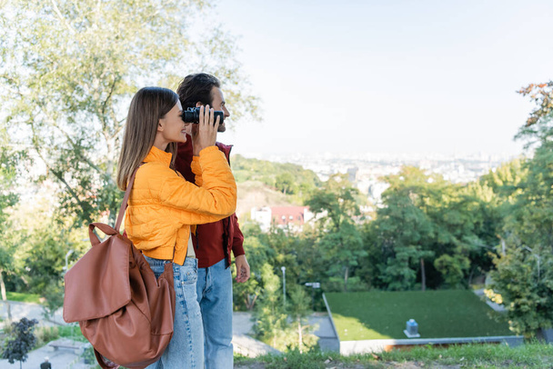 Vista lateral de turista positivo com mochila olhando através de binóculos perto de namorado ao ar livre - Foto, Imagem