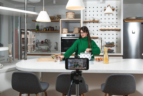 Young vlogger woman vlogging about cooking at her apartment domestic kitchen cook and making video vlog with smartphone as online content creator for internet. Female influencer making a meal. - Zdjęcie, obraz