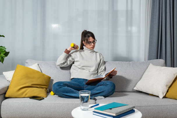 Young happy fitness college student woman sitting at home study reading books to educate herself holding small weight and exercise with one hand. Health and motivated life concept - Foto, Bild