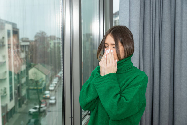 Young sick unhappy woman suffering from common cold or flu standing near window looking out wipes her nose with tissue. Self isolated ill female stay at home until she recover and get well. - Photo, Image