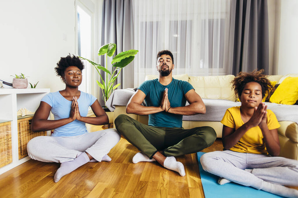 A család jógázik otthon. Lótuszpozícióban vannak, csukott szemmel meditálnak, lélegeznek a nappaliban. - Fotó, kép