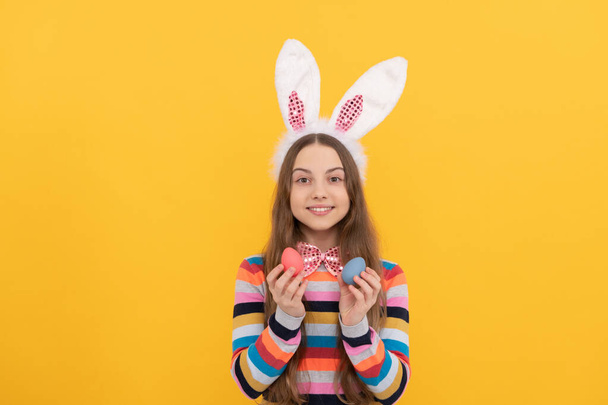 весёлый подросток в кроличьих ушах и галстуке-бабочке держит пасхальные яйца на желтом фоне - Фото, изображение
