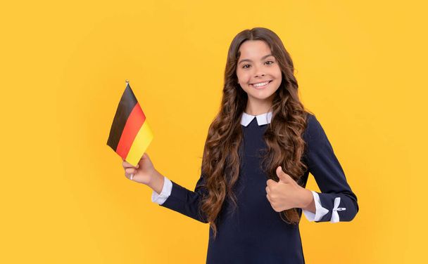 счастливая девочка-подросток держит немецкий флаг на желтом фоне, показывая большой палец вверх, учиться за границей - Фото, изображение
